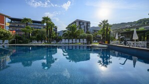 A clear swimming pool surrounded by lounge chairs and umbrellas, with modern buildings and palm trees in the background. - Riviera (Riva del Garda)