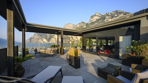 A rooftop terrace with outdoor seating, potted plants, and a view of mountains and the sea. - Riviera (Riva del Garda)