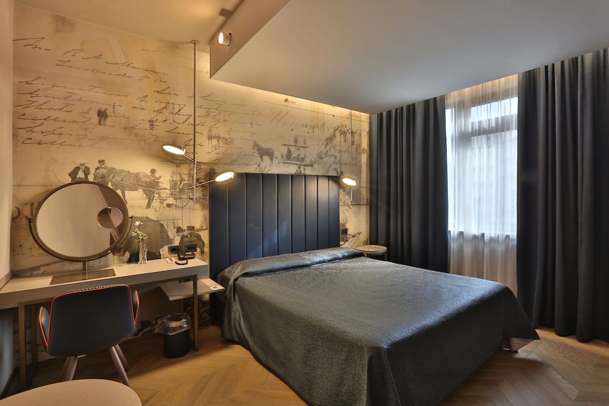 A modern bedroom with a large bed, a desk with a chair, a round mirror, and a wall with historical illustrations.
