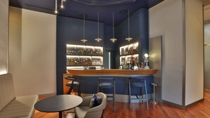 A modern bar area with a wooden counter, bar stools, and a seating area with a sofa and round tables. - Palace Hotel Moderno (Pordenone)