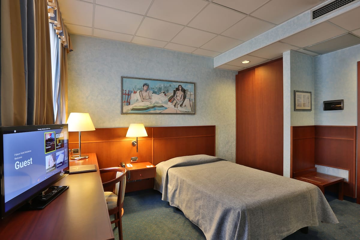 A hotel room with a bed, a desk with a computer, a lamp, a painting on the wall, and a wooden headboard.