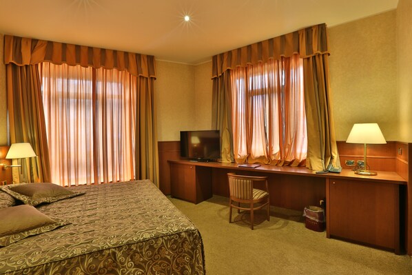 A hotel room with a bed, desk, chair, television, lamps, and curtains.