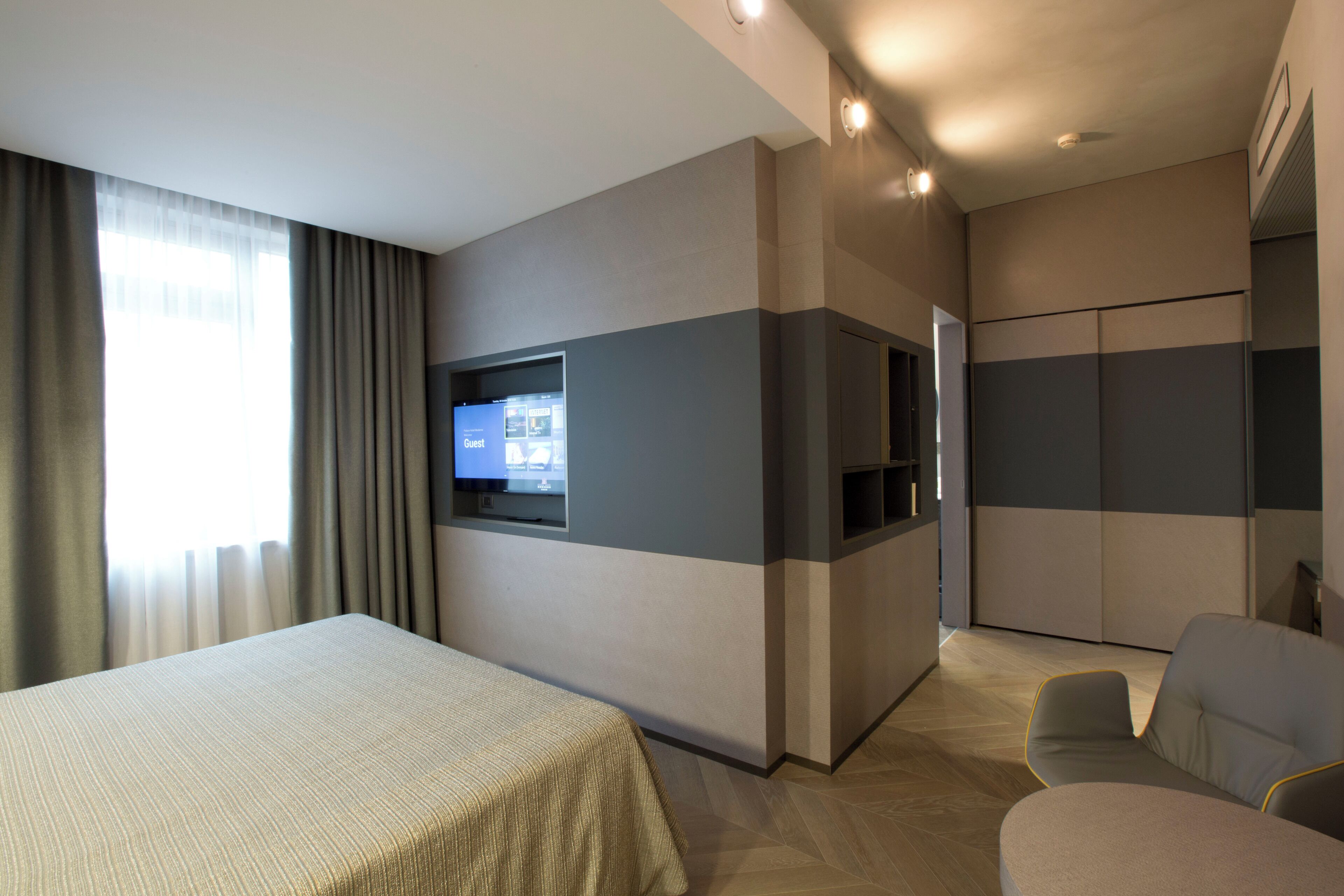 A hotel room with a bed, a TV mounted on the wall, a seating area with chairs, and a wardrobe.