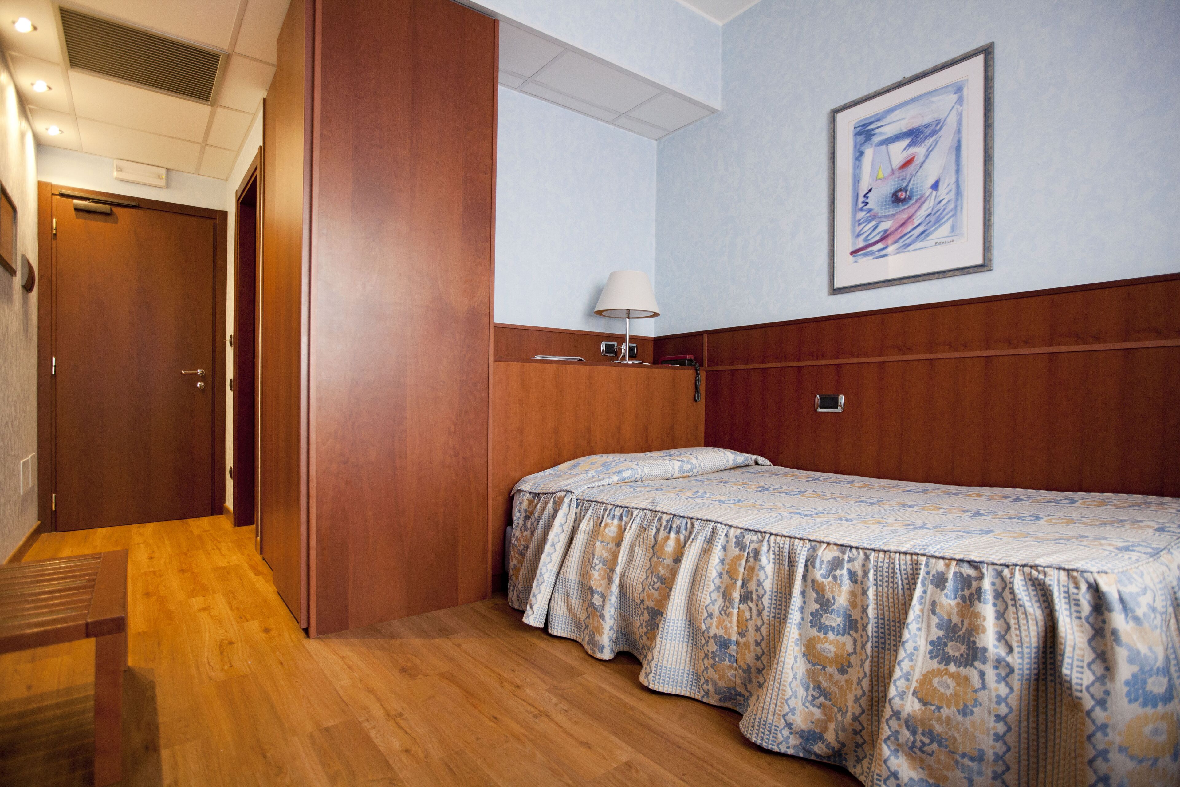 Una habitación de hotel con piso de madera, una cama con cubrecama estampado, una mesita de noche con lámpara, un cuadro enmarcado en la pared y una puerta.