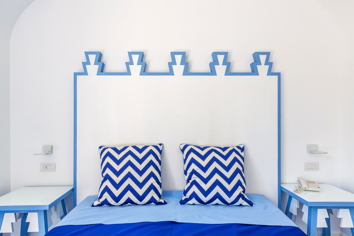 Una cama con cabecera de tablero azul y blanco con estampado en zigzag, dos almohadas a juego y una colcha azul. Hay dos mesitas de noche con tapas blancas y patas azules, cada una con un teléfono blanco y un objeto pequeño encima.