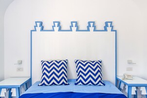Un lit avec une tête de lit à motifs en zigzag bleus et blancs, deux oreillers assortis et une couvre-lit bleue. Il y a deux tables de chevet avec des plateaux blancs et des pieds bleus, chacune surmontée d’un téléphone blanc et d’un petit objet.