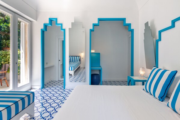 Une chambre d’hôtel moderne dotée d’un grand lit, d’un coin salon avec un banc à rayures et d’une vue sur l’extérieur grâce à une porte vitrée.