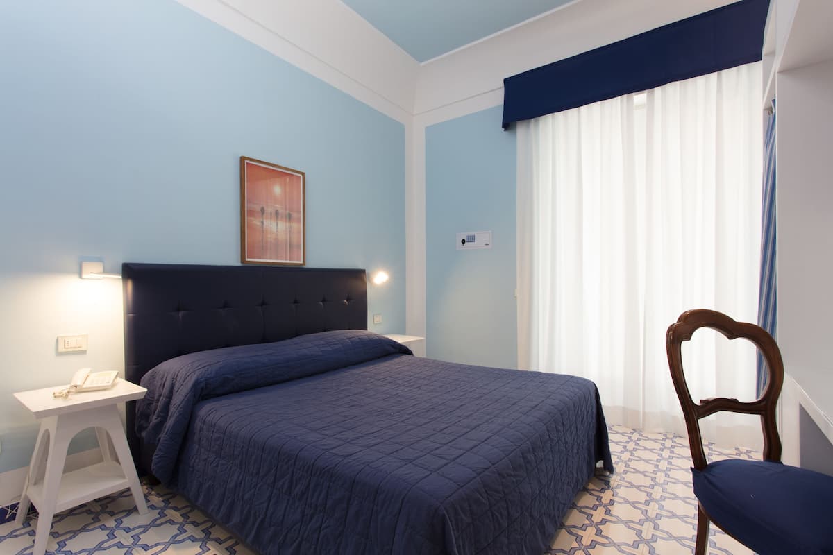 Un dormitorio con cama, una silla, una mesita de noche y una ventana con cortinas.