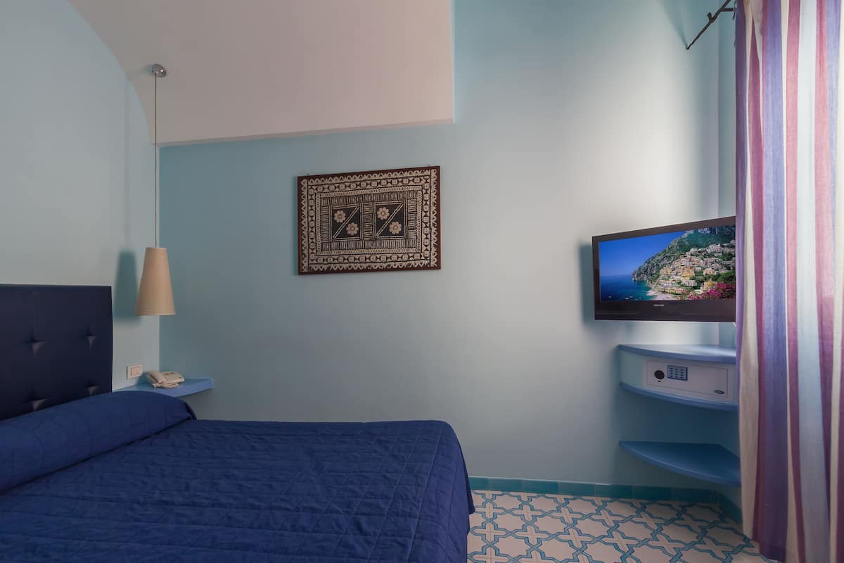 Un dormitorio con una cama, un televisor montado en la pared, una mesita de noche con lámpara y un tapiz decorativo en la pared.