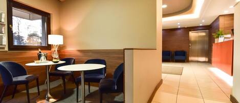 Vestíbulo de hotel con mostrador de recepción, zona de estar con una mesa y sillas, y vista al exterior a través de un ventanal.