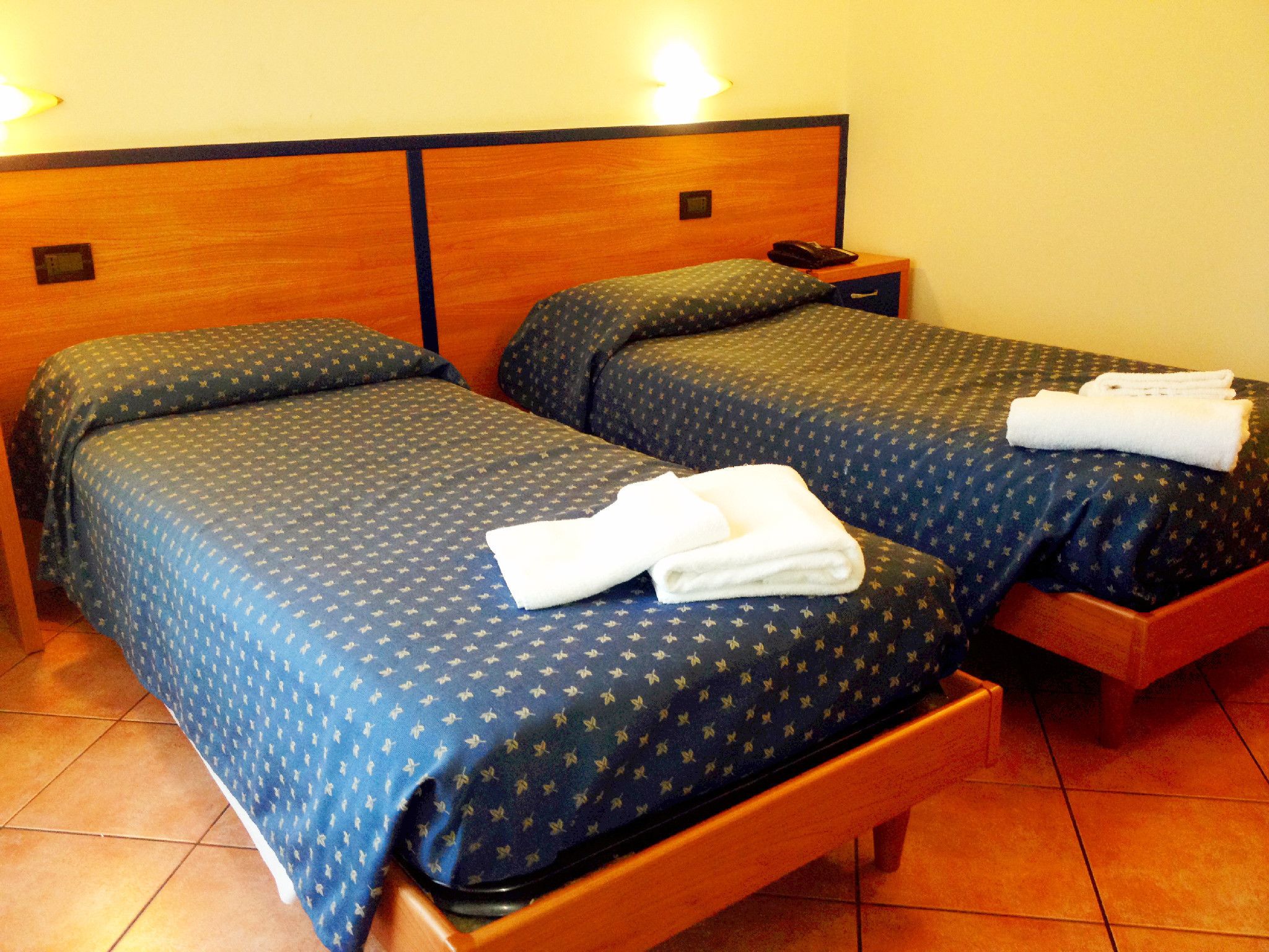 A hotel room with two beds, each with a blue patterned bedspread and pillows, and a wooden headboard. There is a nightstand with a phone and a folded white towel on the bed.