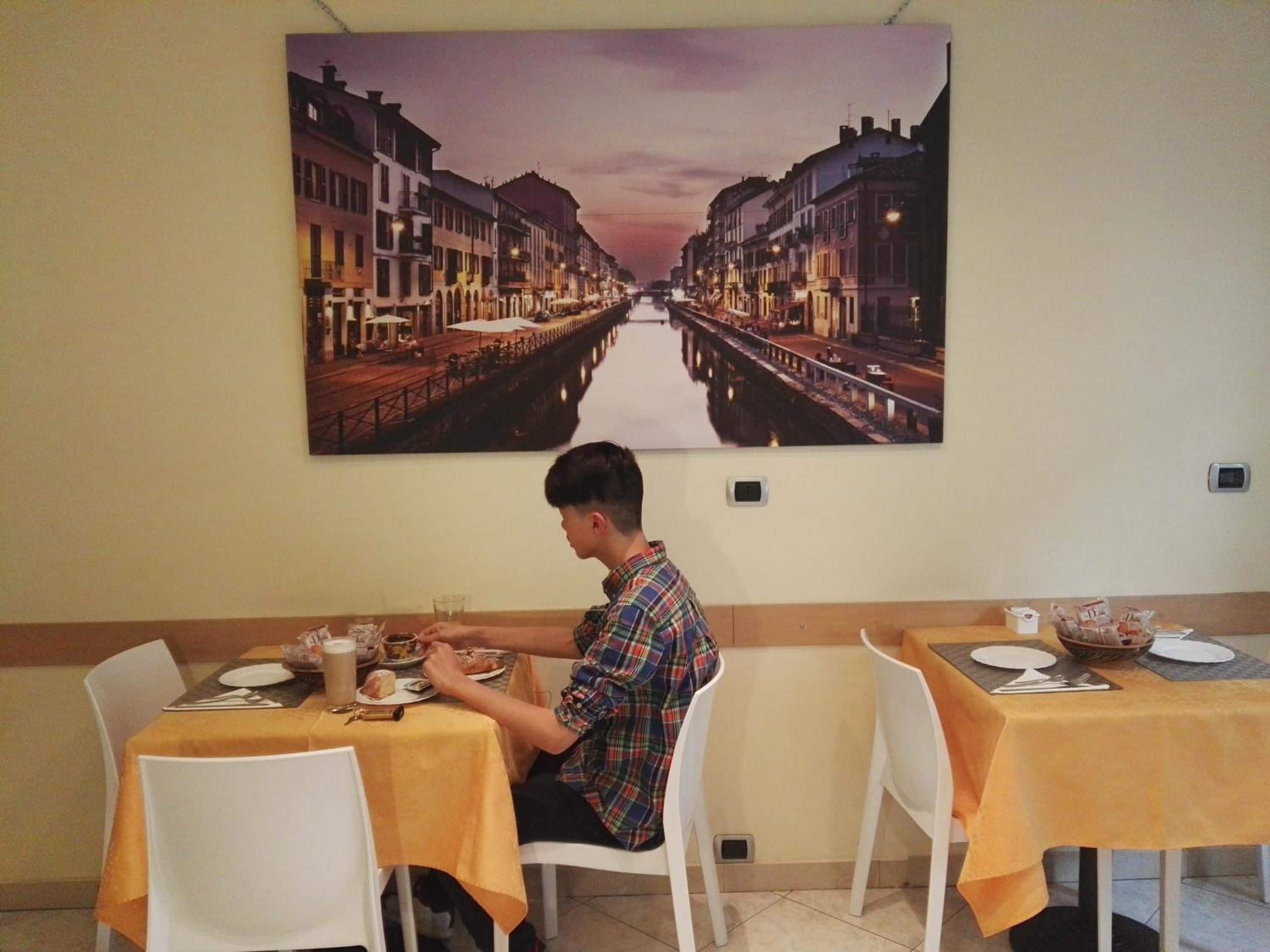 A dining area with tables set for two, a person seated at one table, and a large cityscape painting on the wall.