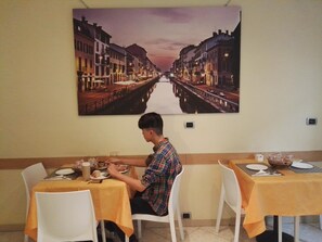 A dining area with tables set for two, a person seated at one table, and a large cityscape painting on the wall. - Hotel Loreto (Milan)
