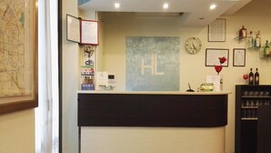 A hotel reception desk with a sign reading "HL" and a clock above it. There are framed documents and a map on the wall, and a shelf with bottles and glasses to the right. - Hotel Loreto (Milan)