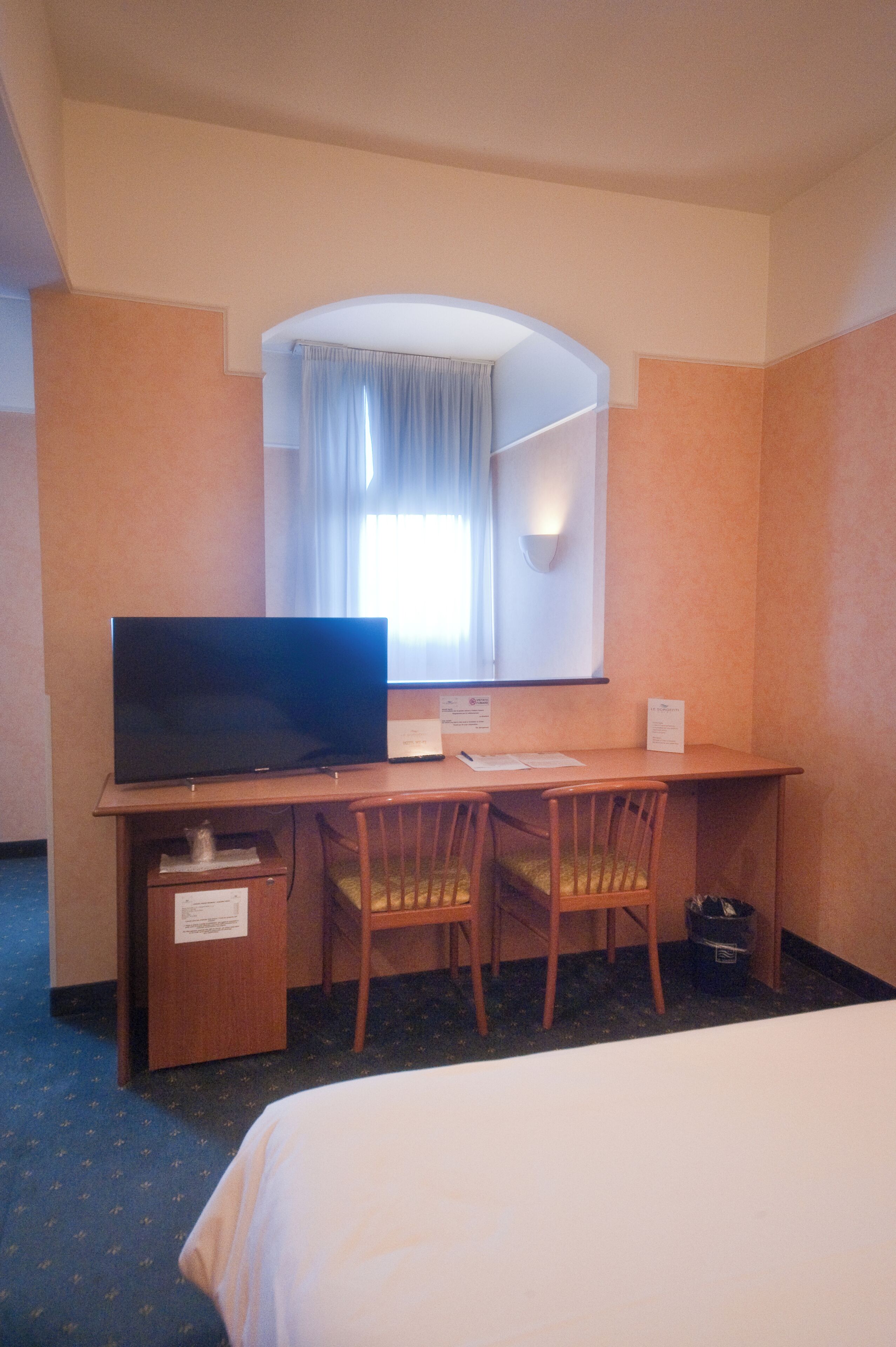 A hotel room with a bed, a desk with a television, two chairs, a window with curtains, and a wall-mounted light.