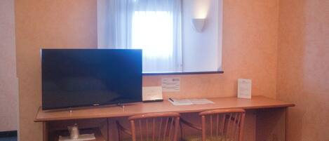 A hotel room with a bed, a desk with a television, two chairs, a window with curtains, and a wall-mounted light.