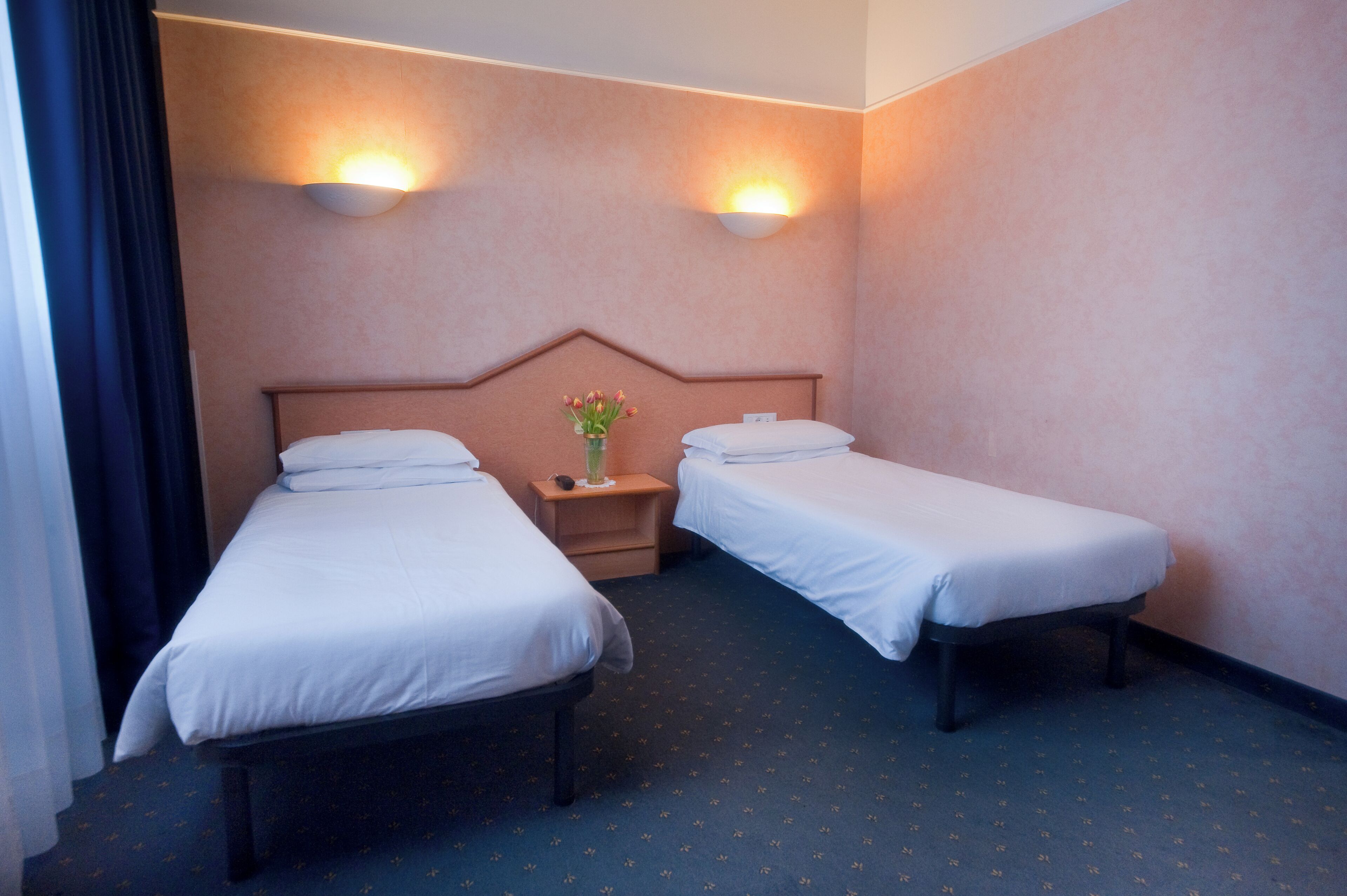 A hotel room with two single beds, a nightstand with a vase of flowers, and wall-mounted lights.