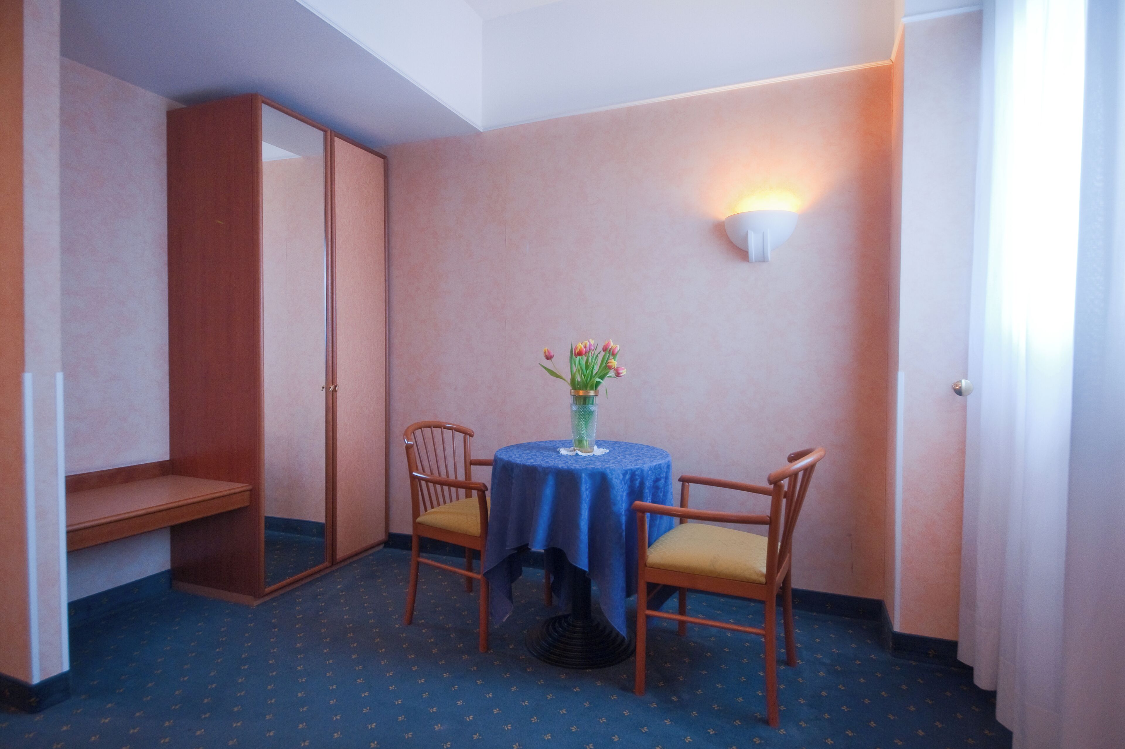 Ein kleines Zimmer mit einem runden Tisch, zwei Stühlen, einer Blumenvase, einem Kleiderschrank und einer Wandleuchte.