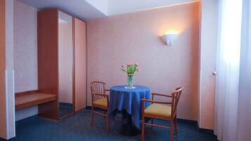 Ein kleines Zimmer mit einem runden Tisch, zwei Stühlen, einer Blumenvase, einem Kleiderschrank und einer Wandleuchte.