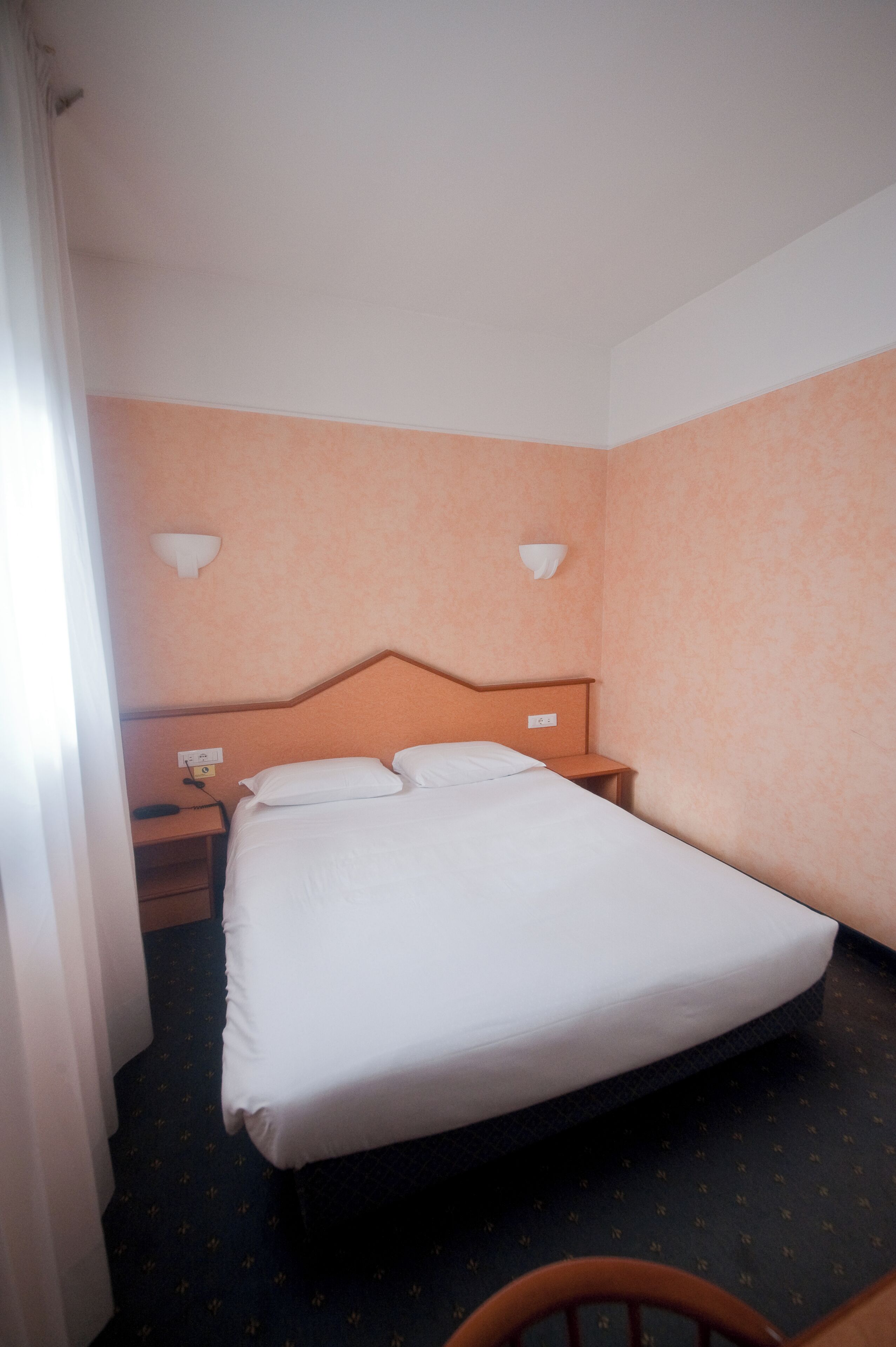 A hotel room with a single bed, a nightstand, two wall-mounted lamps, and a power outlet.