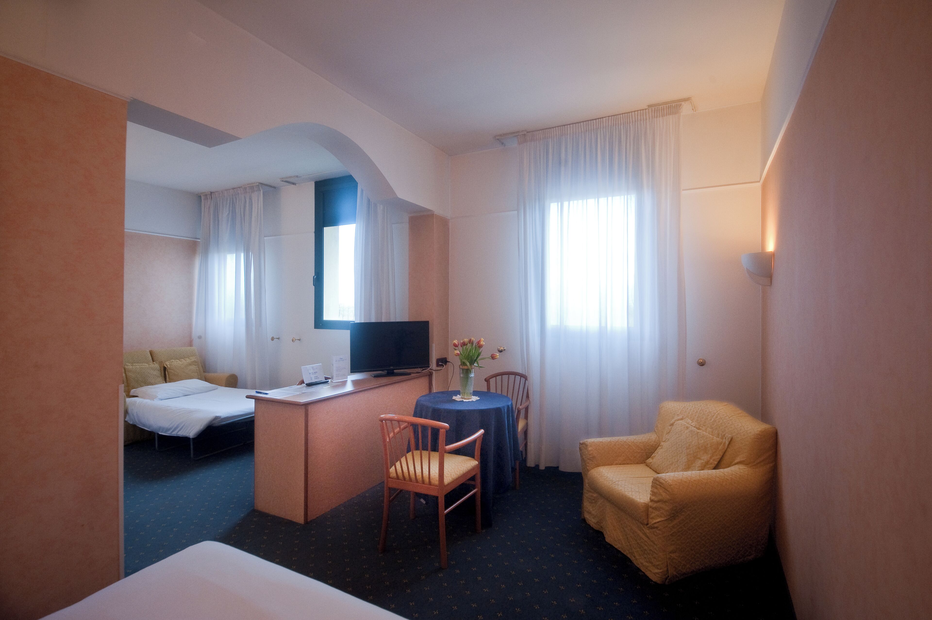A hotel room with a bed, a desk with a chair, a television, and a small table with a vase of flowers.