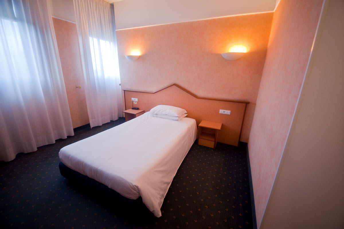 Habitación de hotel con una cama individual, una mesita de noche, dos lámparas fijadas a la pared y un ventanal con cortinas traslúcidas.