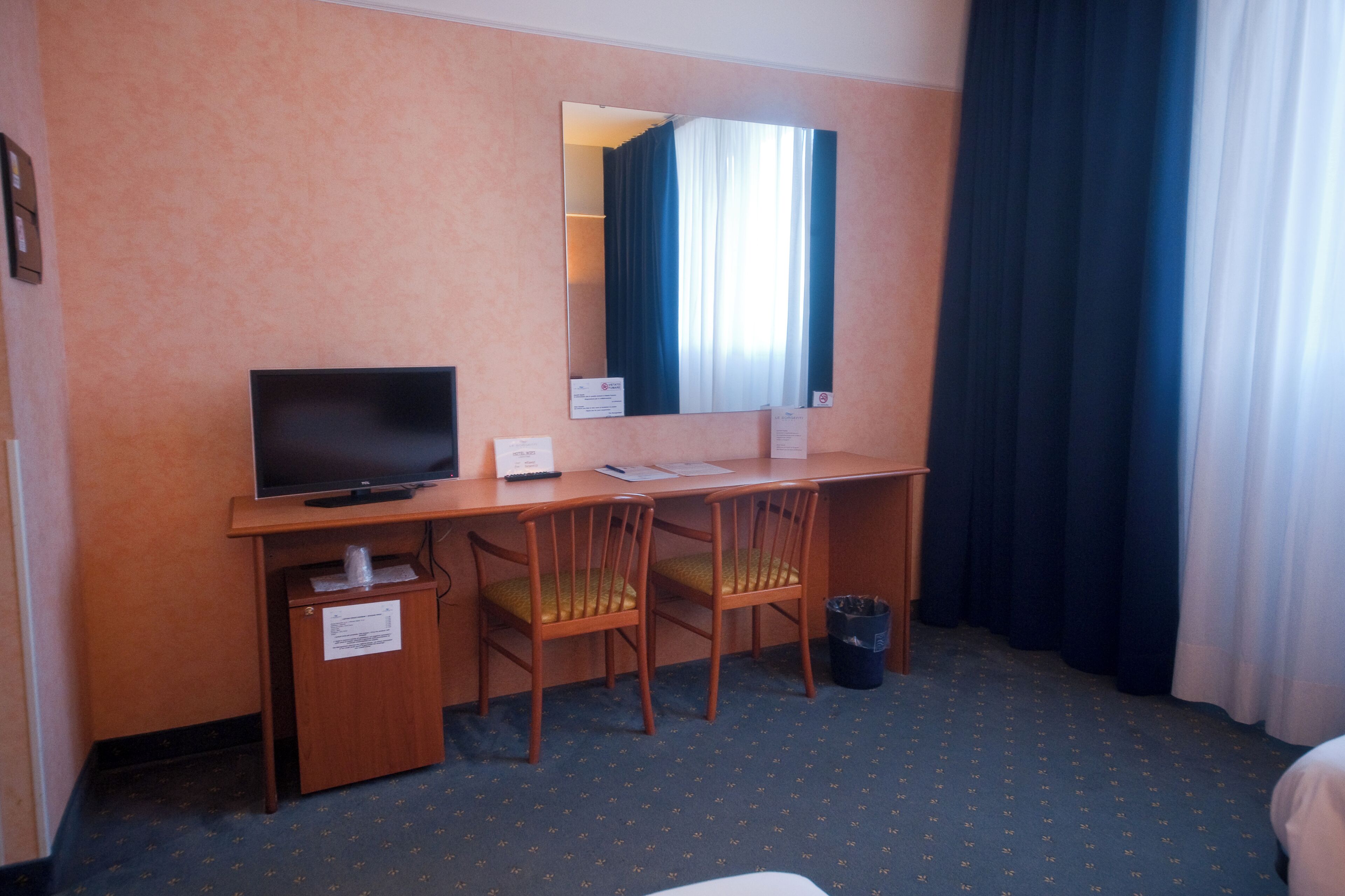 Una camera d'albergo con una scrivania, due sedie, una televisione, uno specchio e una finestra con tende.