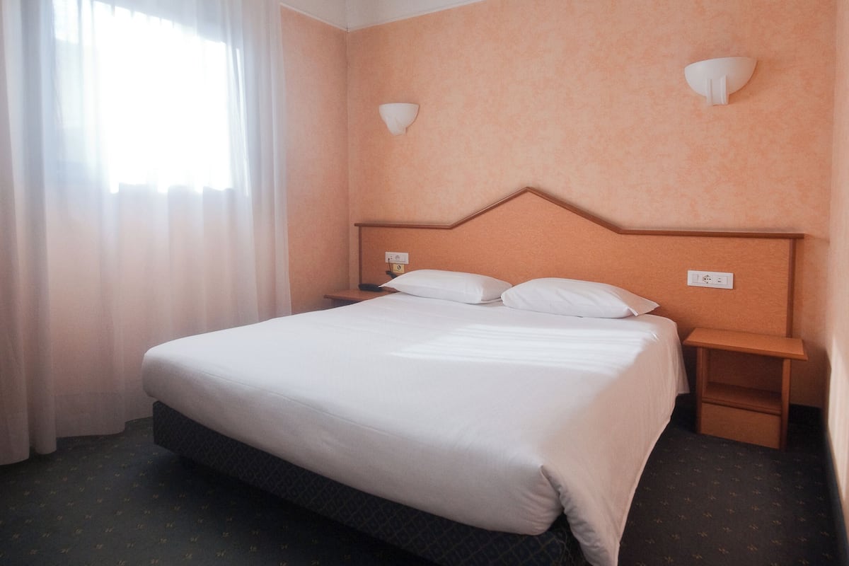 Una habitación de hotel con una cama grande, una mesita de noche, una lámpara de pared y una ventana con cortinas translúcidas.