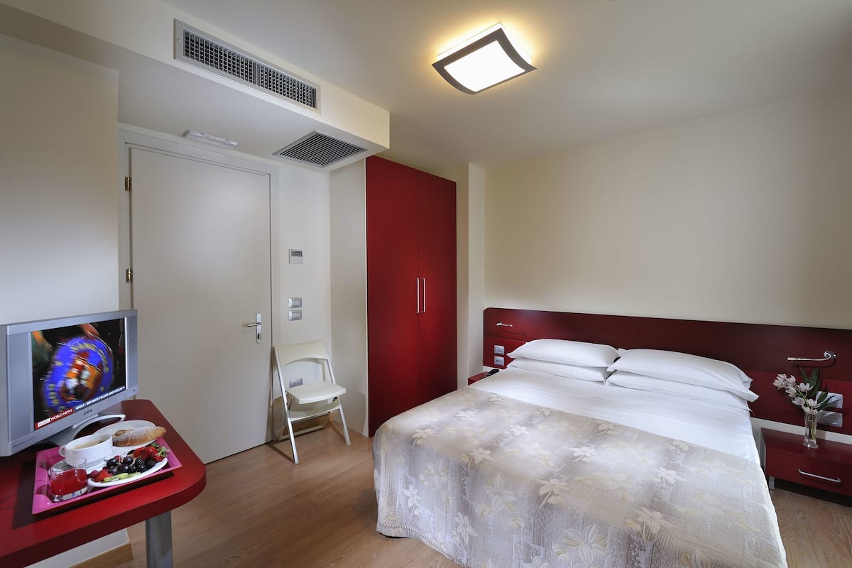 Hotellihuone, jossa on sänky, televisio, työpöytä, jonka päällä on tarjotinalus ruokaa varten, sekä punainen vaatekaappi.
