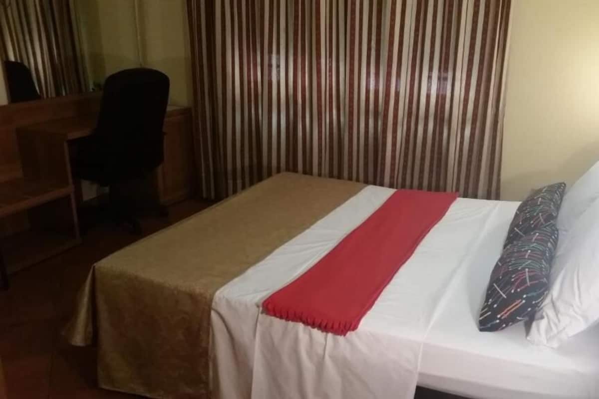 Un lit simple avec une couvre-lit rouge et blanche, un oreiller à motifs multicolores et un bureau en bois avec une chaise.