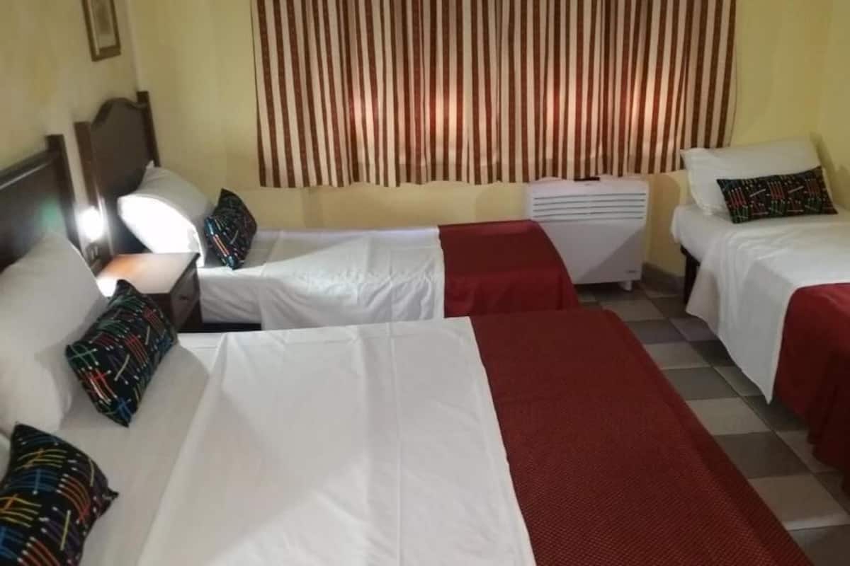 Une chambre d’hôtel avec deux lits simples, une petite table, une chaise, un radiateur et une fenêtre avec des stores verticaux.