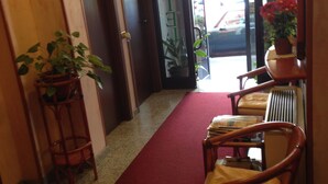 Una hall d'albergo con tappeto rosso, sedie in legno, una pianta in vaso e una porta con la scritta 'HOTEL'.