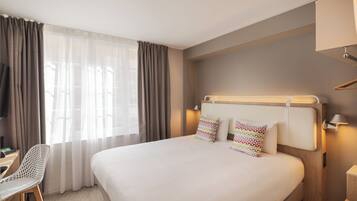 A hotel room with a large bed, two pillows, a desk with a chair, and a window with curtains.