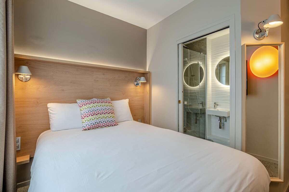A hotel room with a large bed, two bedside lamps, a wooden headboard, a glass shower enclosure, and a round mirror.