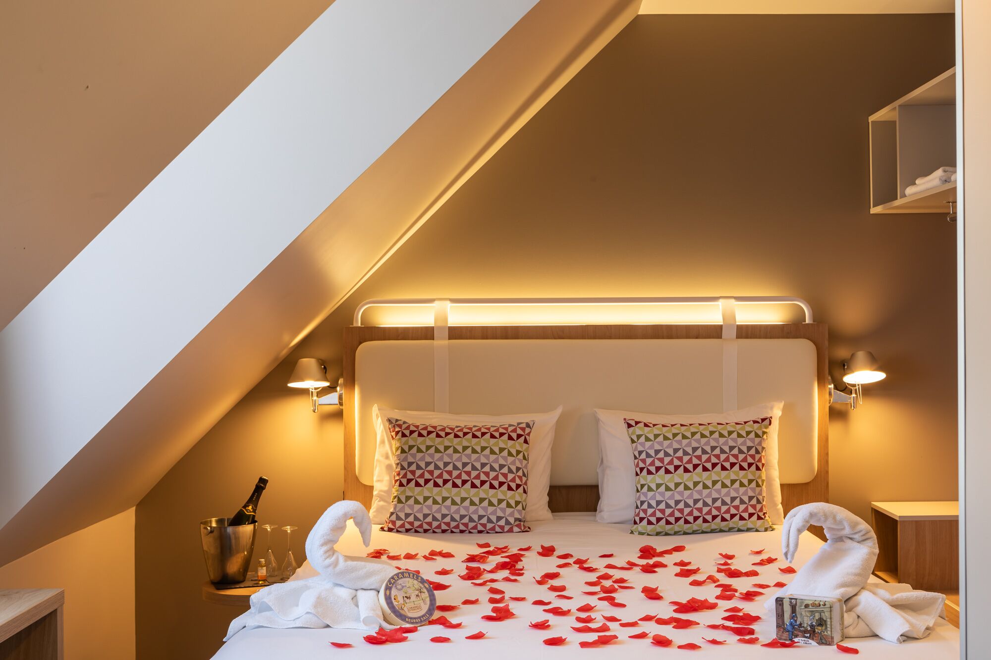 A hotel room with a bed, two patterned pillows, a bottle of champagne, and a swan-shaped towel.
