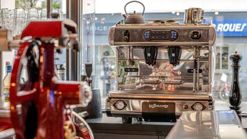 Een koffieapparaat met een digitaal display en stoomkraan, geplaatst op een toonbank met een glazen vitrine vol bekers op de achtergrond.