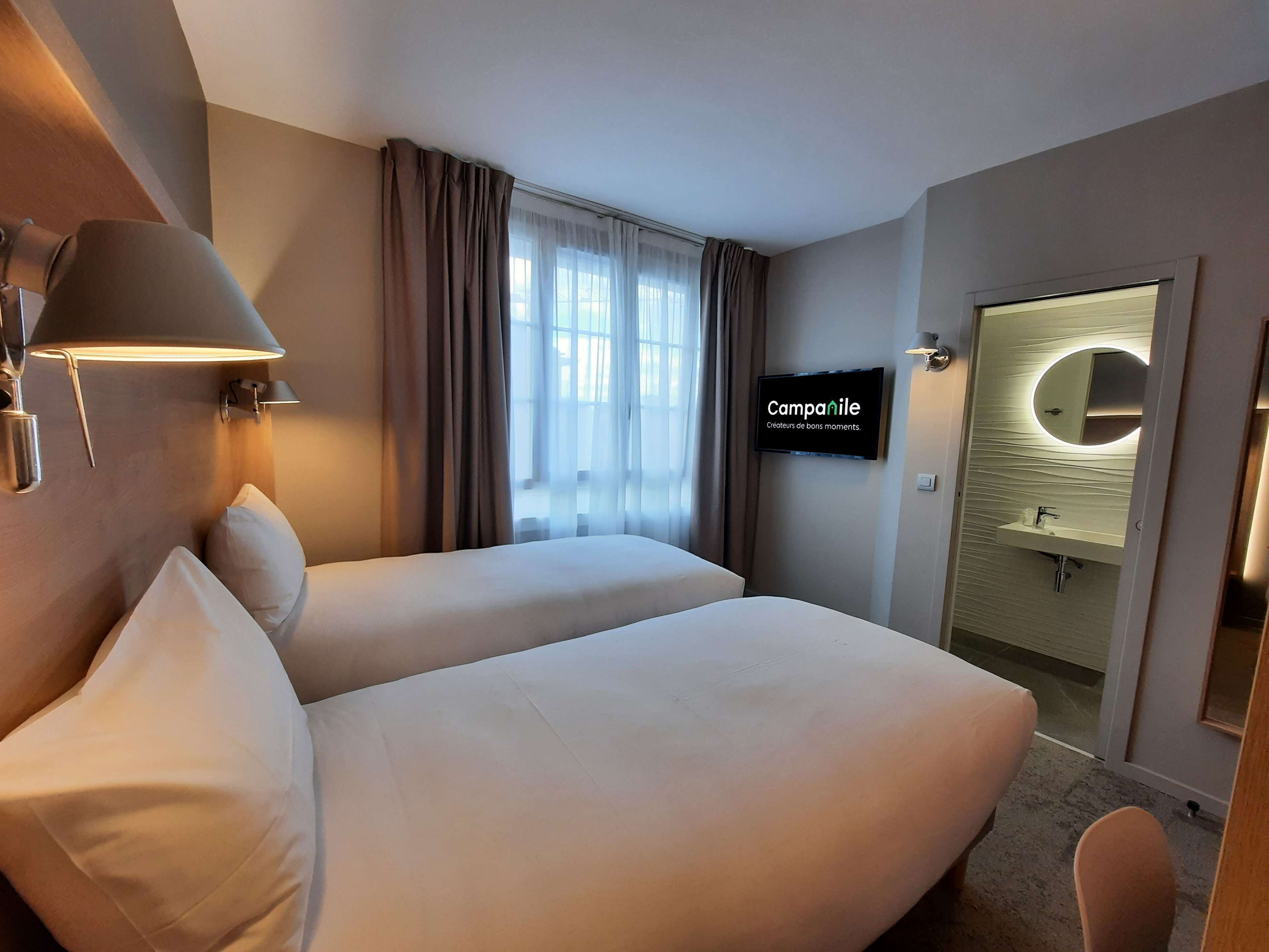 A hotel room with two beds, a TV mounted on the wall, a bathroom with a mirror and sink, and a window with curtains.