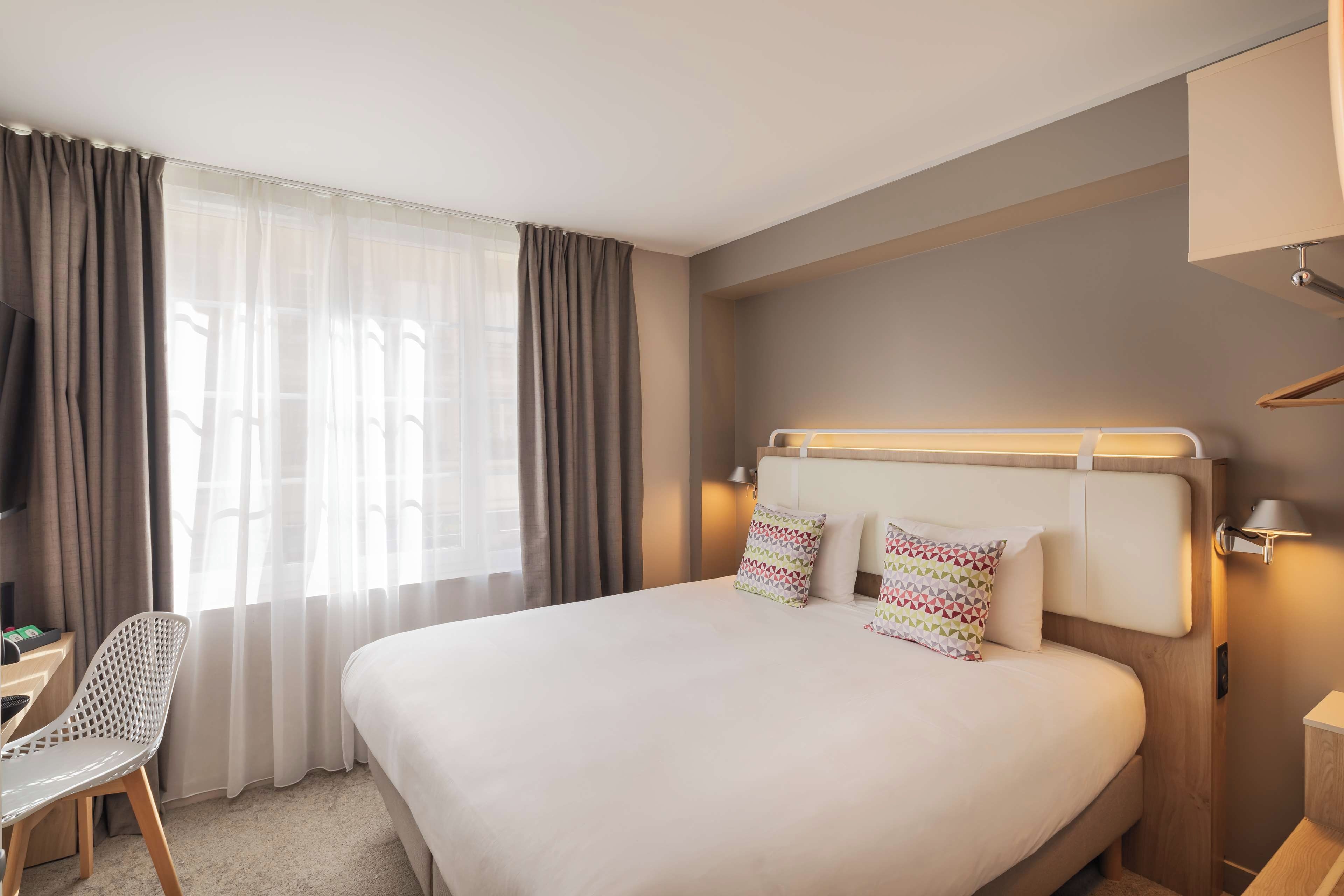 A hotel room with a bed, a desk, a chair, and a window with curtains.
