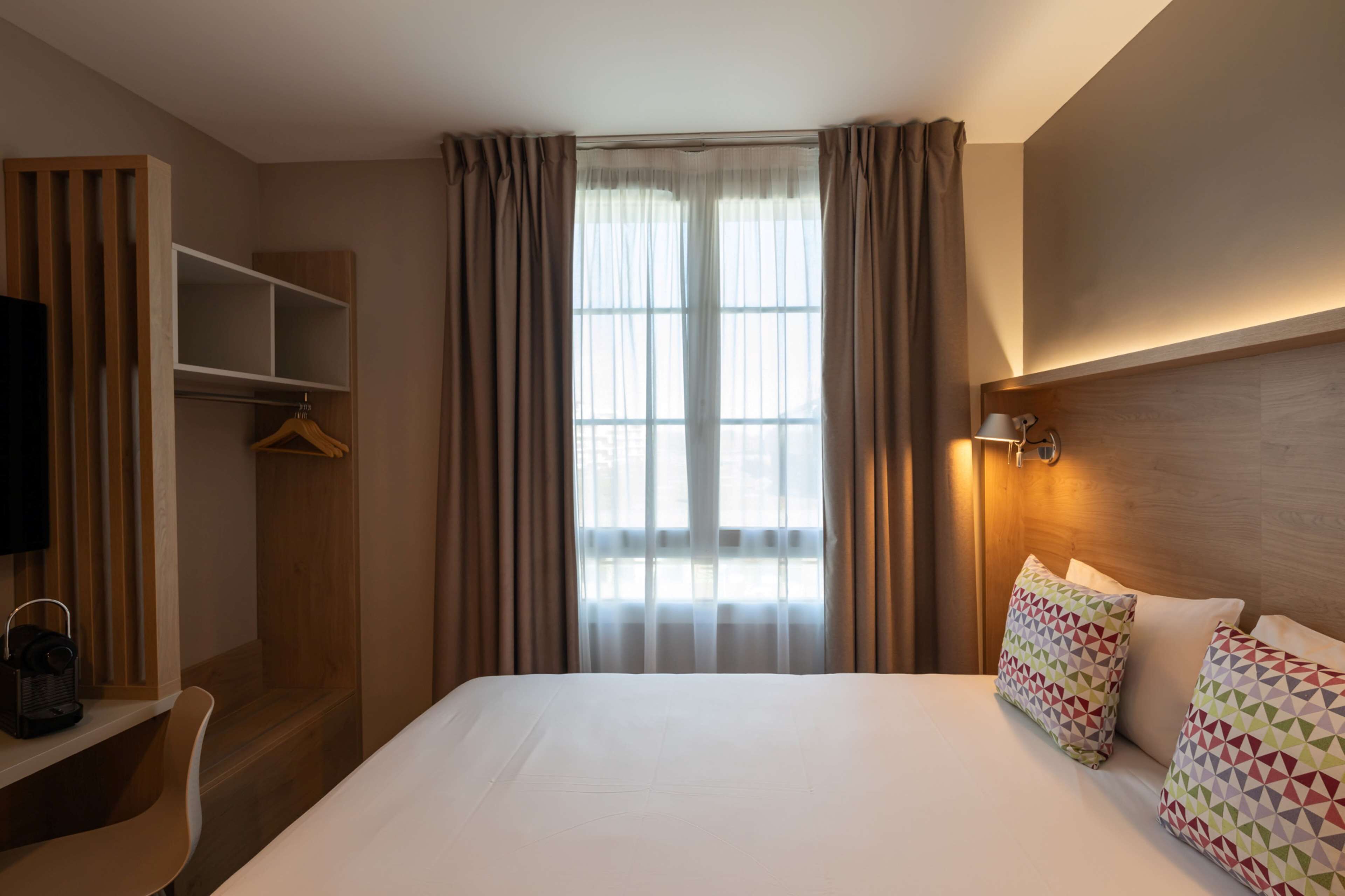A hotel room with a bed, a desk with a chair, a TV, a wardrobe, and a window with curtains.
