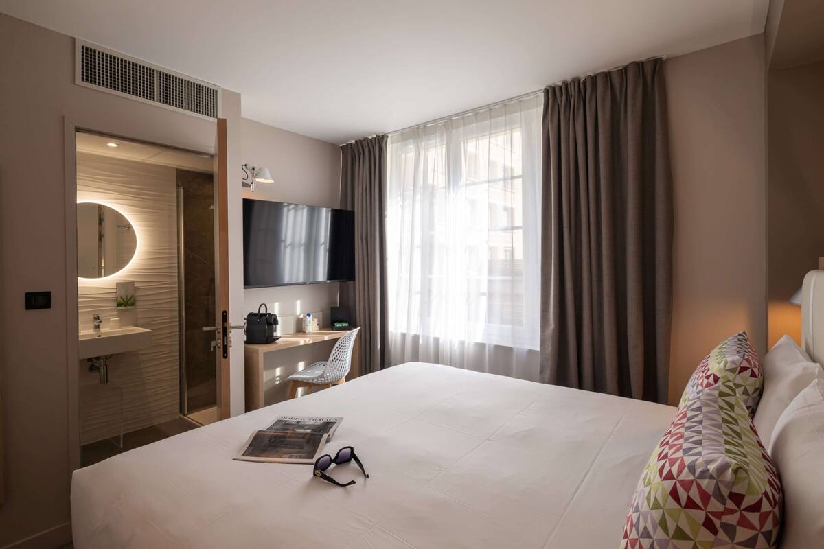 Habitación de hotel con cama, escritorio, silla, televisor, baño con espejo y ventana con cortinas.