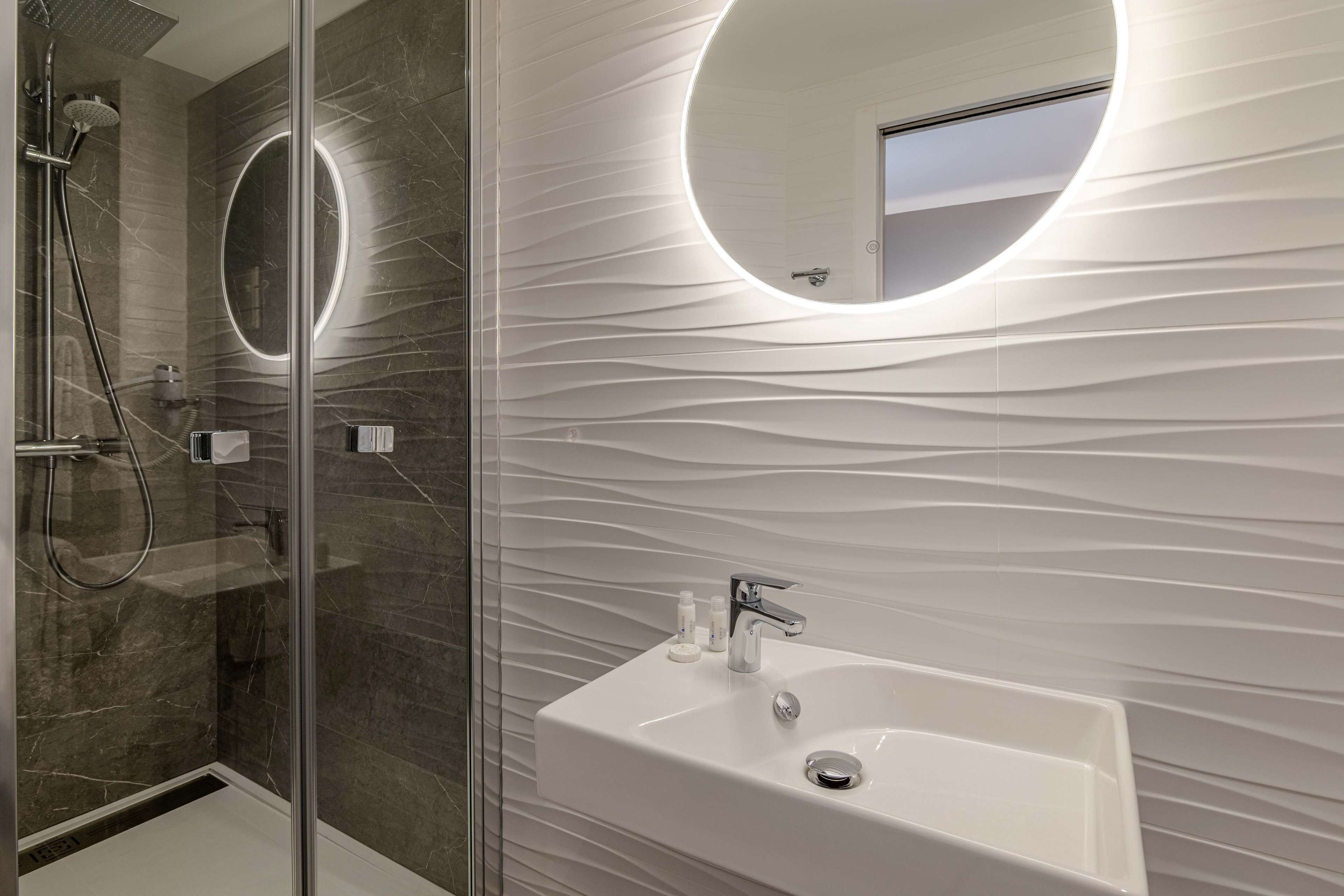 Un baño moderno con ducha de vidrio, un lavabo blanco con un solo grifo y un espejo adosado a la pared.