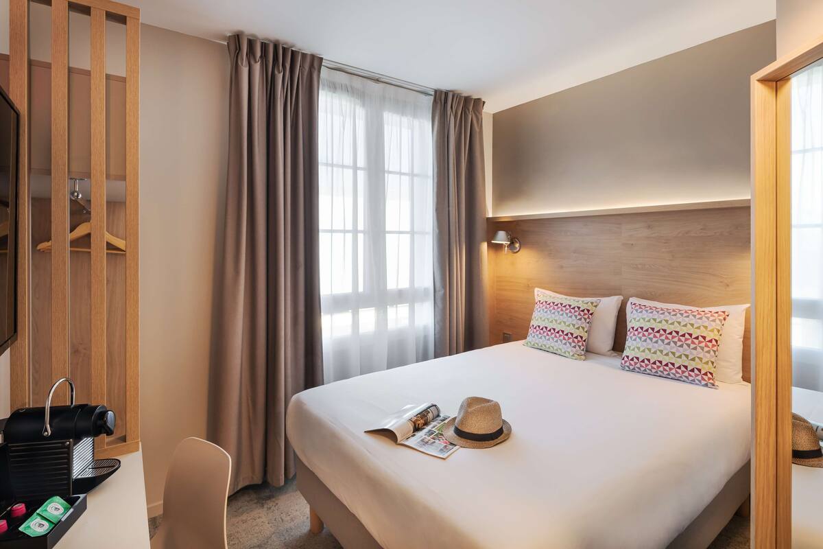Una habitación de hotel con cama, escritorio con cafetera, perchero y ventana con cortinas.