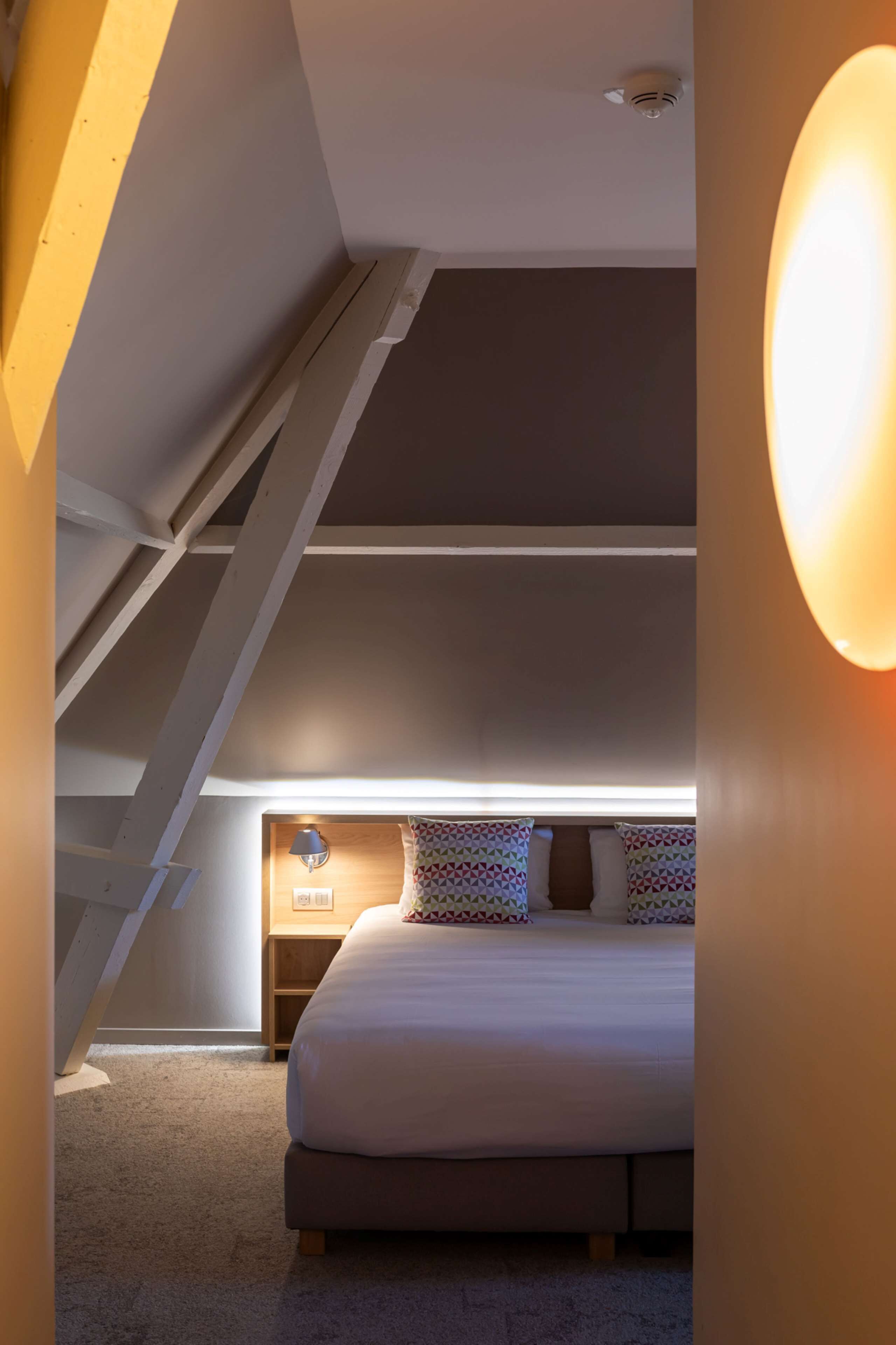 A bedroom with a sloped ceiling, a bed with patterned pillows, a bedside lamp, and a small wooden nightstand.