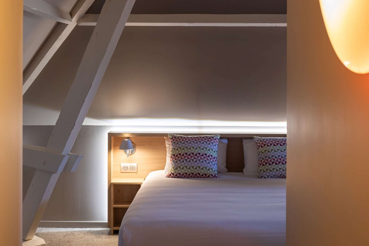 A bedroom with a sloped ceiling, a bed with patterned pillows, a bedside lamp, and a small wooden nightstand.