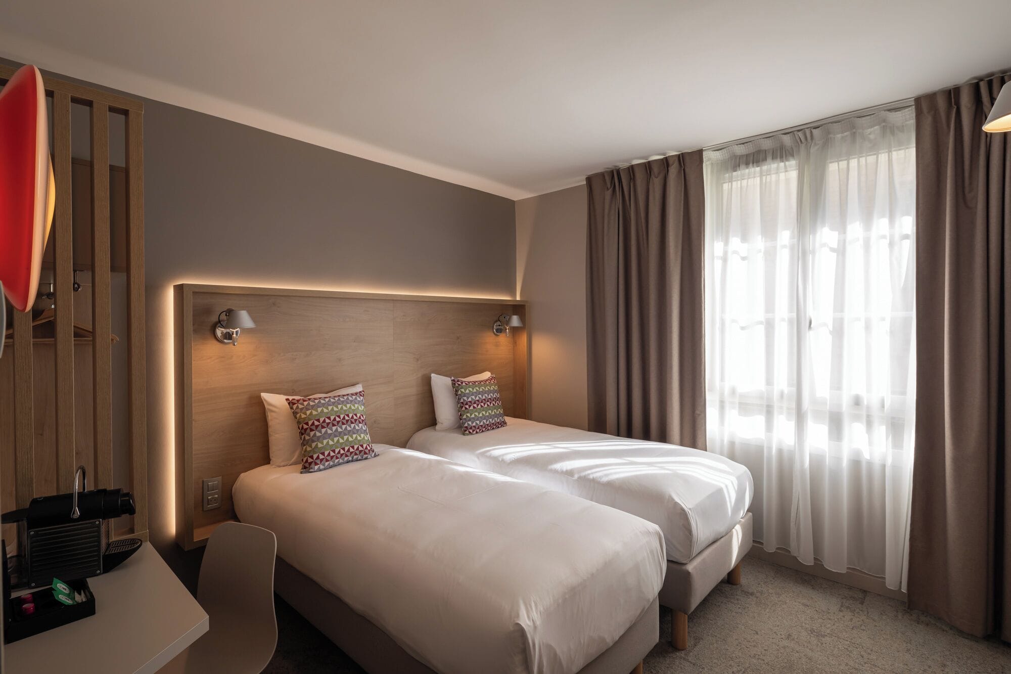 Habitación de hotel con dos camas, cabecero de madera, escritorio con silla y ventana con cortinas.