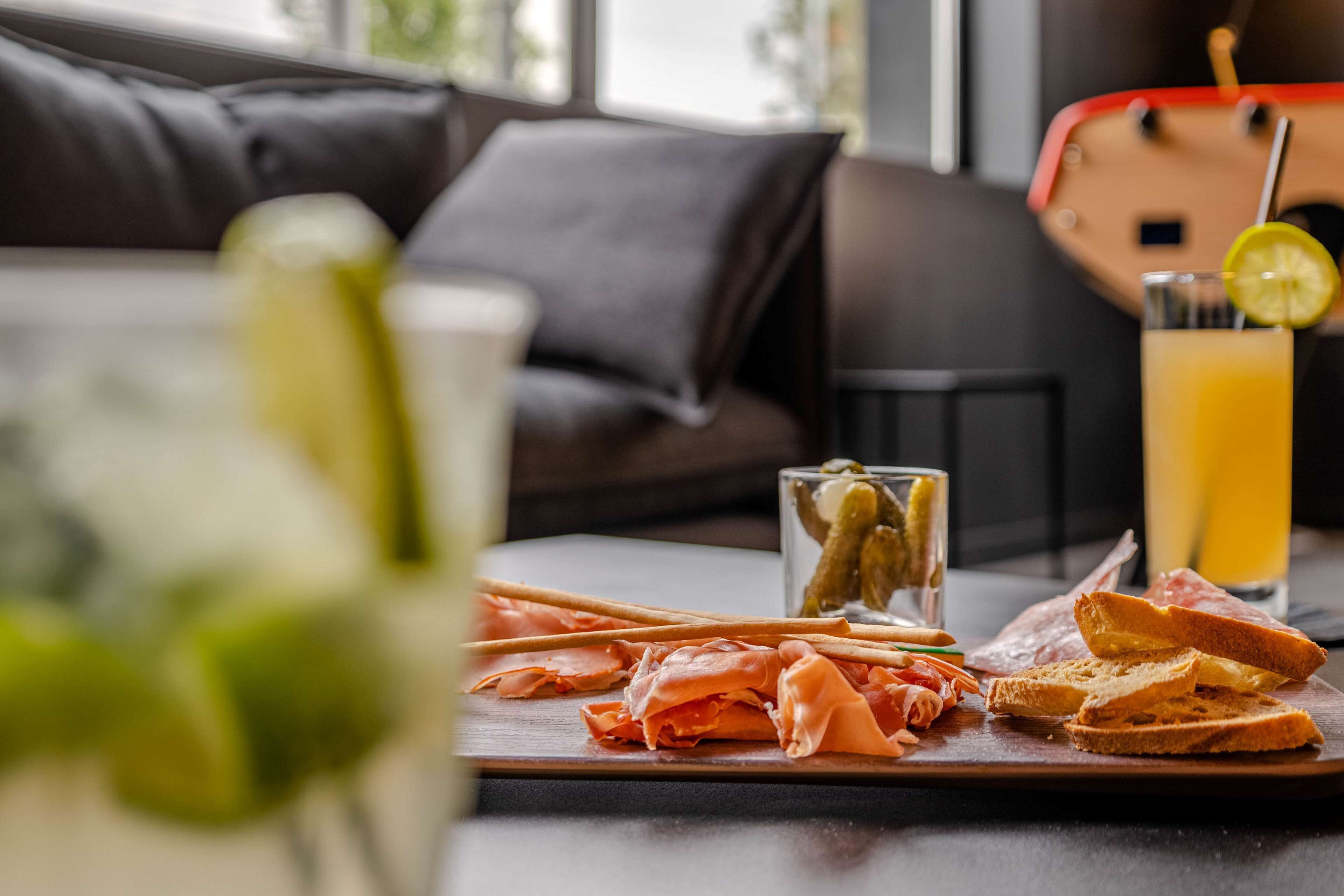 A dining table with a wooden board holding sliced meats, a glass of juice, and a glass with a lemon slice.