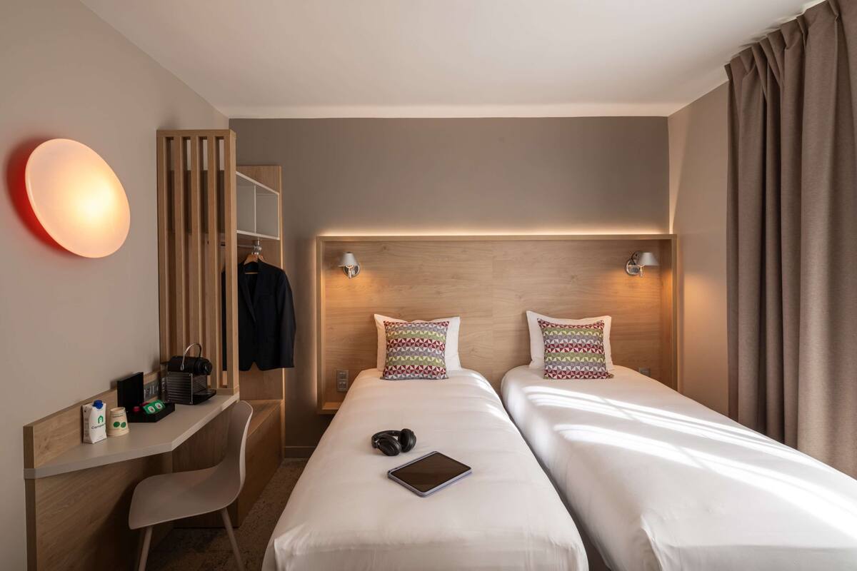 Habitación de hotel con dos camas, un escritorio con silla, perchero y lámpara de pared.