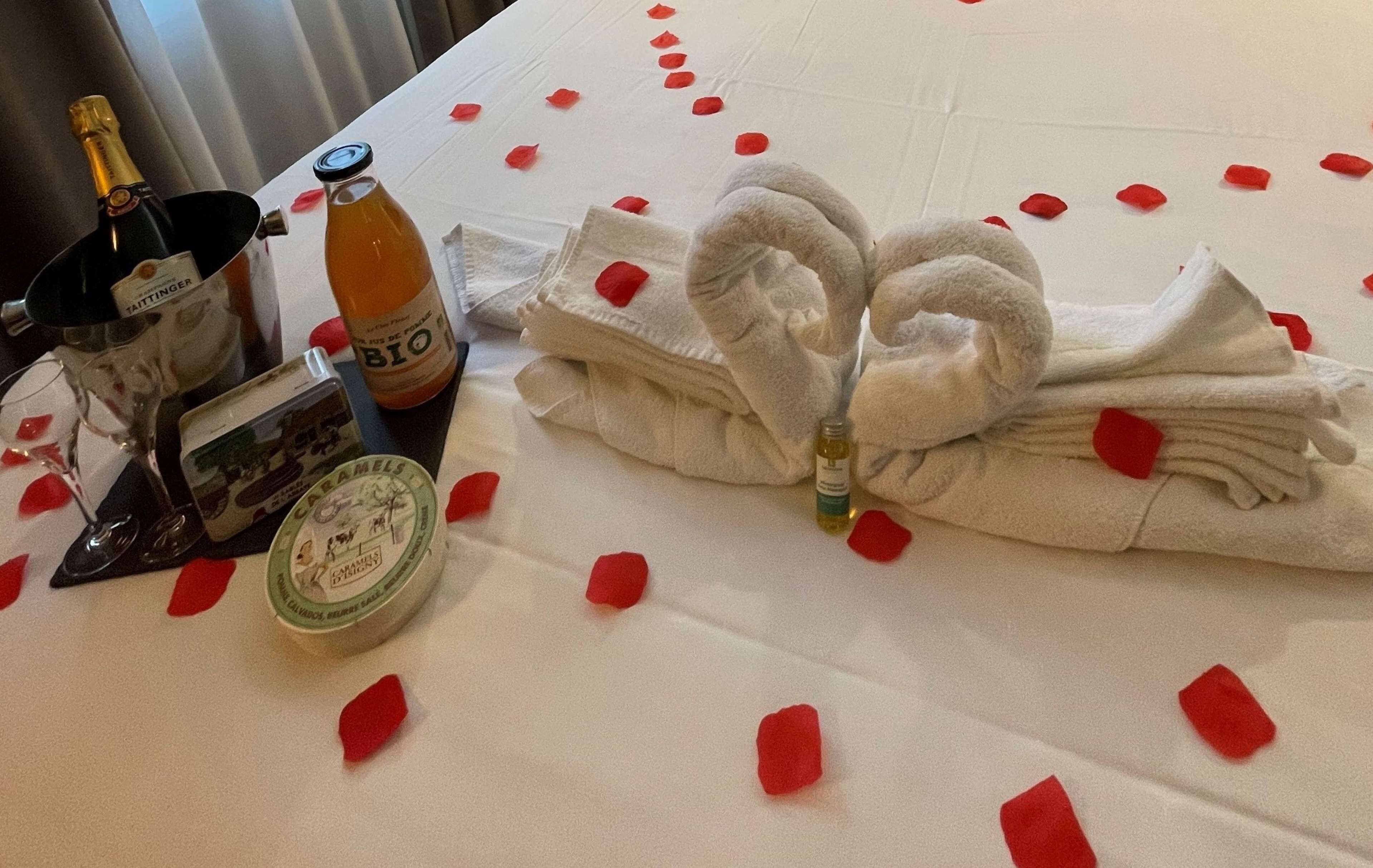 A neatly made bed with rose petals, a bottle of BIO, a bottle of champagne, and a small gift box.