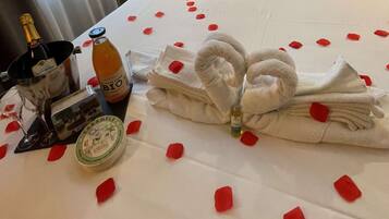A neatly made bed with rose petals, a bottle of BIO, a bottle of champagne, and a small gift box.