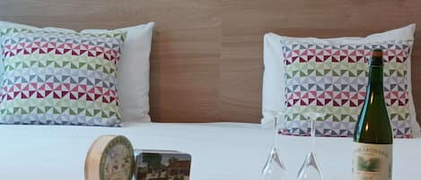 Habitación de hotel con cabecera de madera, almohadas estampadas, una botella de vino, dos copas y un pequeño objeto decorativo sobre la cama.